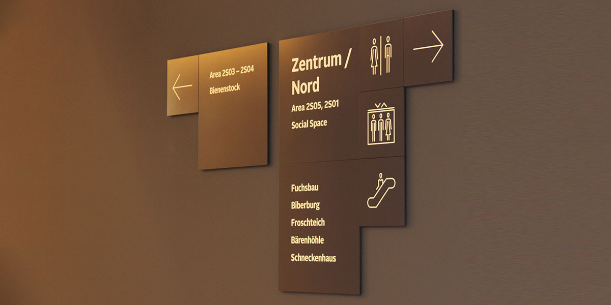 Bronzefarbene Wandschilder mit Richtungspfeilen, Bereichsnamen und Piktogrammen für WC, Aufzug und Rolltreppe