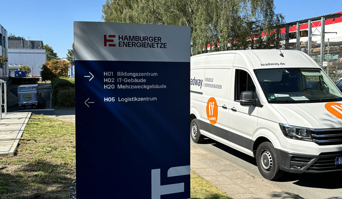 Monolith mit Wegweisern zu Bildungszentrum, IT-Gebäude, Mehrzweckgebäude und Logistikzentrum vor einem Firmengelände, daneben ein Montagefahrzeug von Leadway