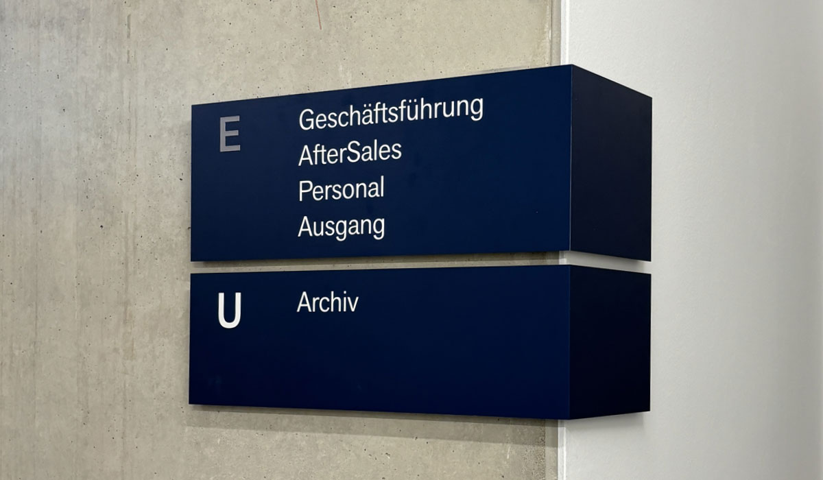 Zweiteiliges dunkelblaues Wandschild mit den Etagenkennungen E und U und den Zielen Geschäftsführung, AfterSales, Personal, Ausgang und Archiv