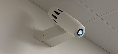 Weißer Lichtprojektor an einem Wandausleger unter einer Rasterdecke, das Objektiv leuchtet blau