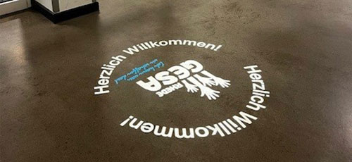 Auf einen Betonboden projiziertes GESA-Logo, kreisförmig umrahmt vom Schriftzug Herzlich Willkommen