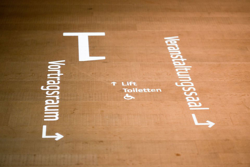 Lichtprojektion auf einem Holzboden mit Pfeilen zu Vortragsraum, Veranstaltungssaal, Lift und barrierefreien Toiletten
