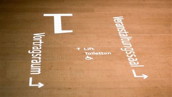 Lichtprojektion auf einem Holzboden mit Pfeilen zu Vortragsraum, Veranstaltungssaal, Lift und barrierefreien Toiletten