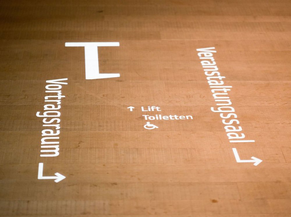 Lichtprojektion auf einem Holzboden mit Pfeilen zu Vortragsraum, Veranstaltungssaal, Lift und barrierefreien Toiletten