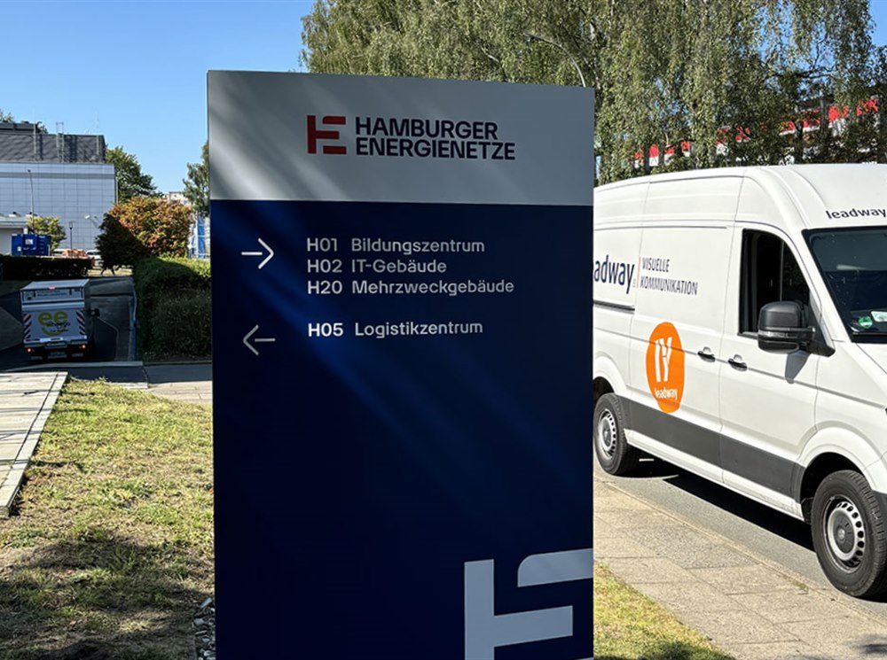 Monolith mit Wegweisern zu Bildungszentrum, IT-Gebäude und Logistikzentrum vor einem Firmengelände, daneben ein Montagefahrzeug von Leadway