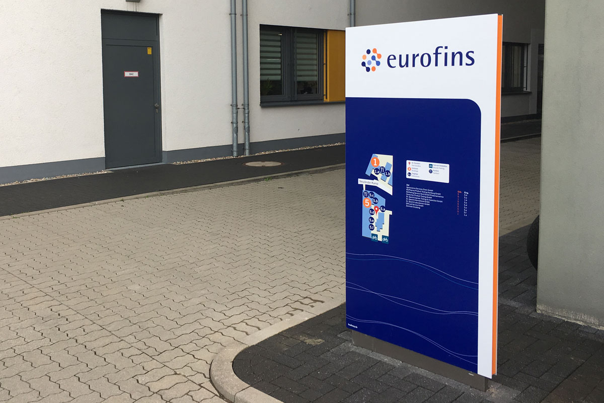 Weißblauer Monolith mit Eurofins-Logo, Lageplan des Geländes und Firmenverzeichnis vor einem Bürogebäude
