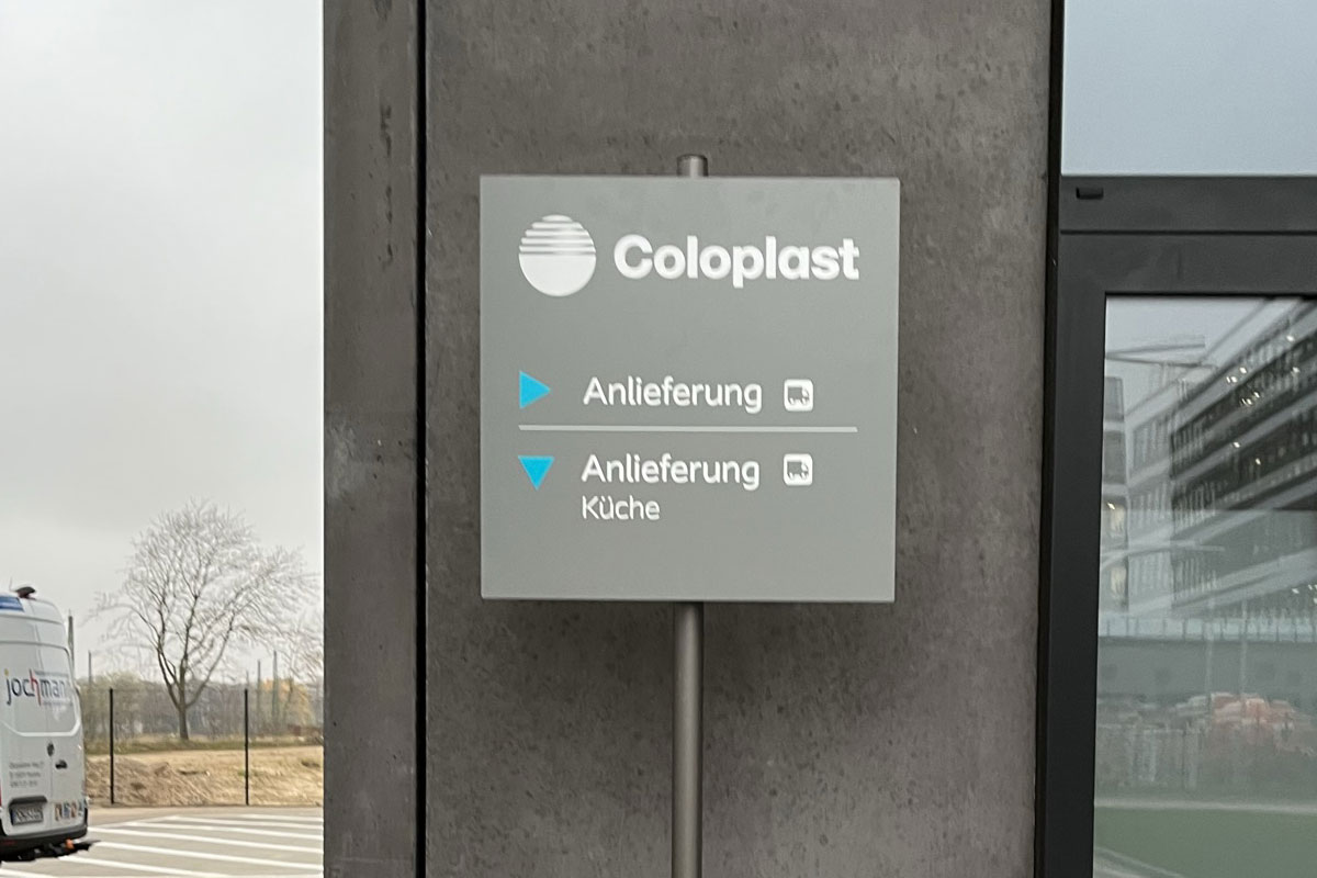 Freistehendes graues Hinweisschild auf einem Pfosten mit Coloplast-Logo und zwei Wegweisern zur Anlieferung mit Lkw-Piktogrammen