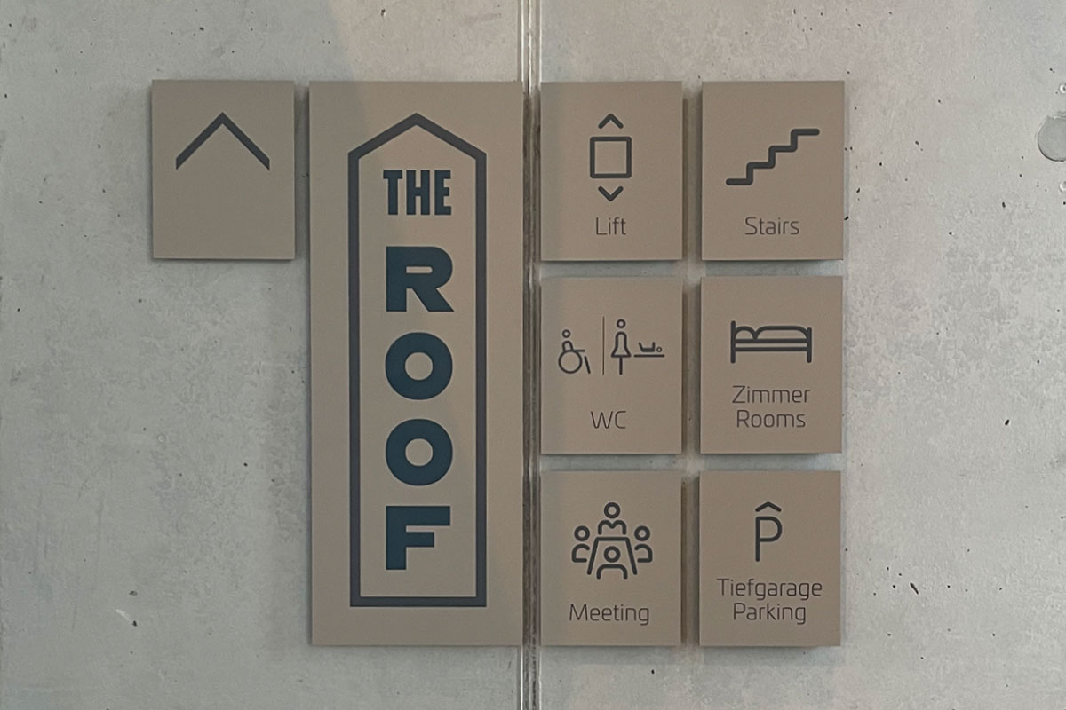 Beigefarbene Wandschilder auf Sichtbeton mit dem Schriftzug THE ROOF und Piktogrammfeldern für Lift, Treppe, WC, Zimmer, Meeting und Tiefgarage