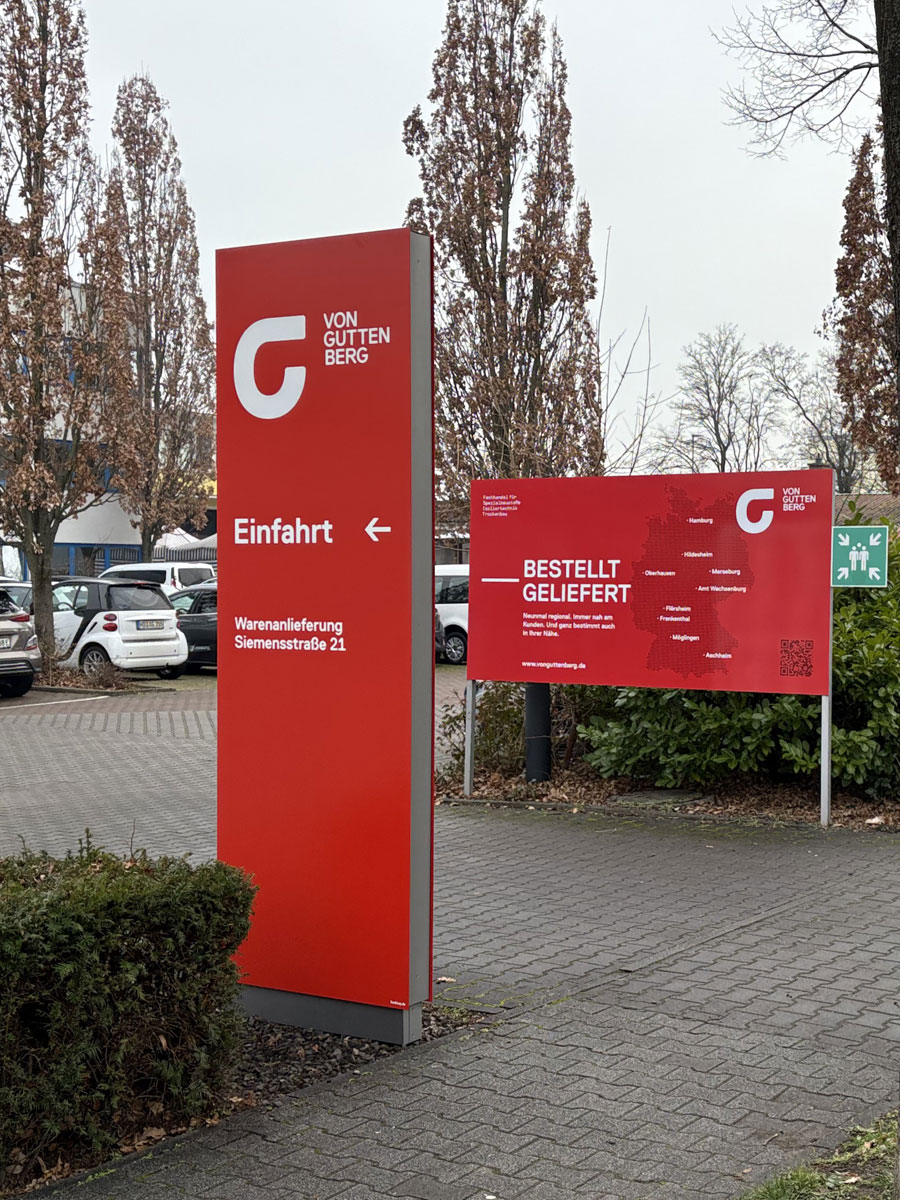 Roter Monolith von von Guttenberg mit Einfahrtspfeil und Adresse der Warenanlieferung, daneben eine großformatige Werbetafel mit Deutschlandkarte