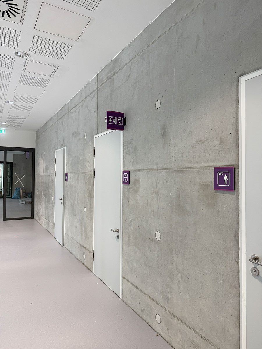 Flur mit Sichtbetonwänden und violetten WC-Schildern als Wand- und Flügelschilder neben weißen Türen