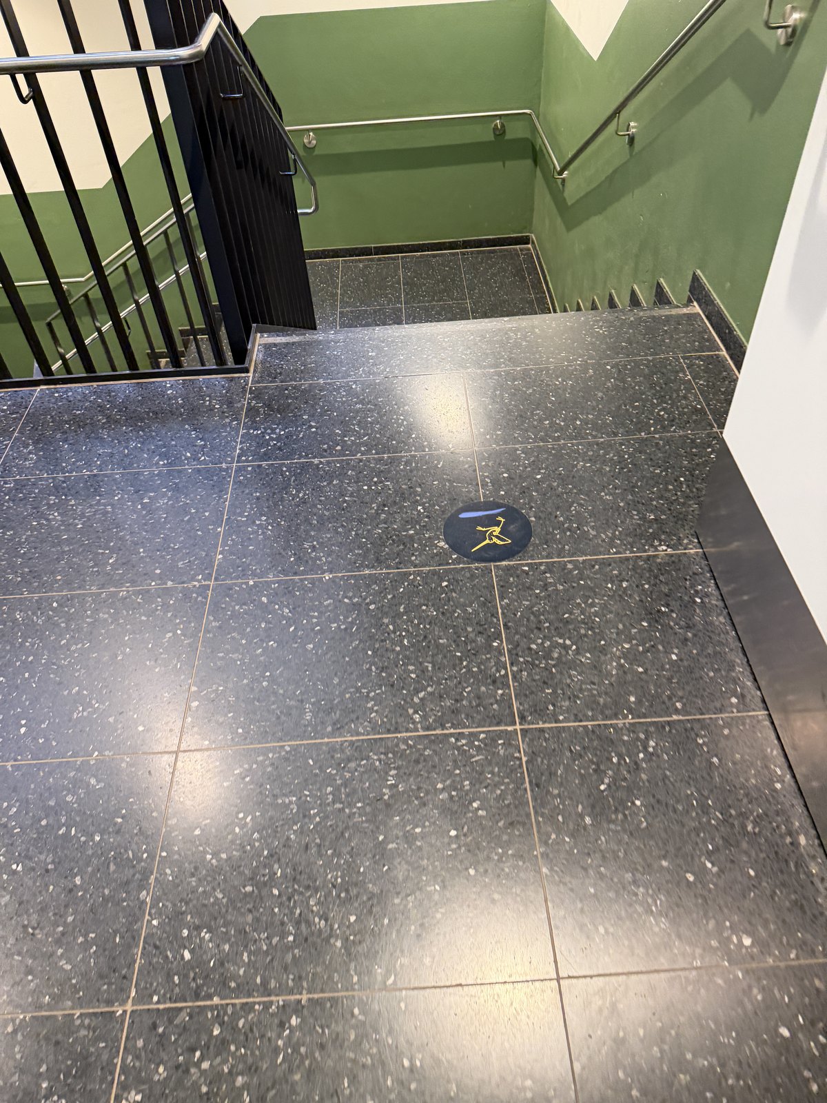 Dunkler Terrazzo-Boden an einem Treppenpodest mit einer runden Bodenfolie: eine gelbe Bekassine als Linienzeichnung über einem blauen Pinselstrich