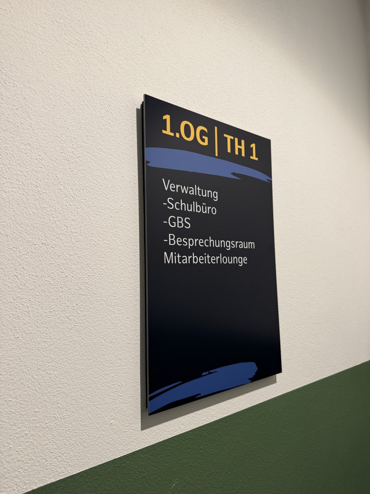 Wegweiser mit der gelben Überschrift 1.OG | TH 1 und den Zielen Verwaltung, Schulbüro, GBS, Besprechungsraum, Mitarbeiterlounge