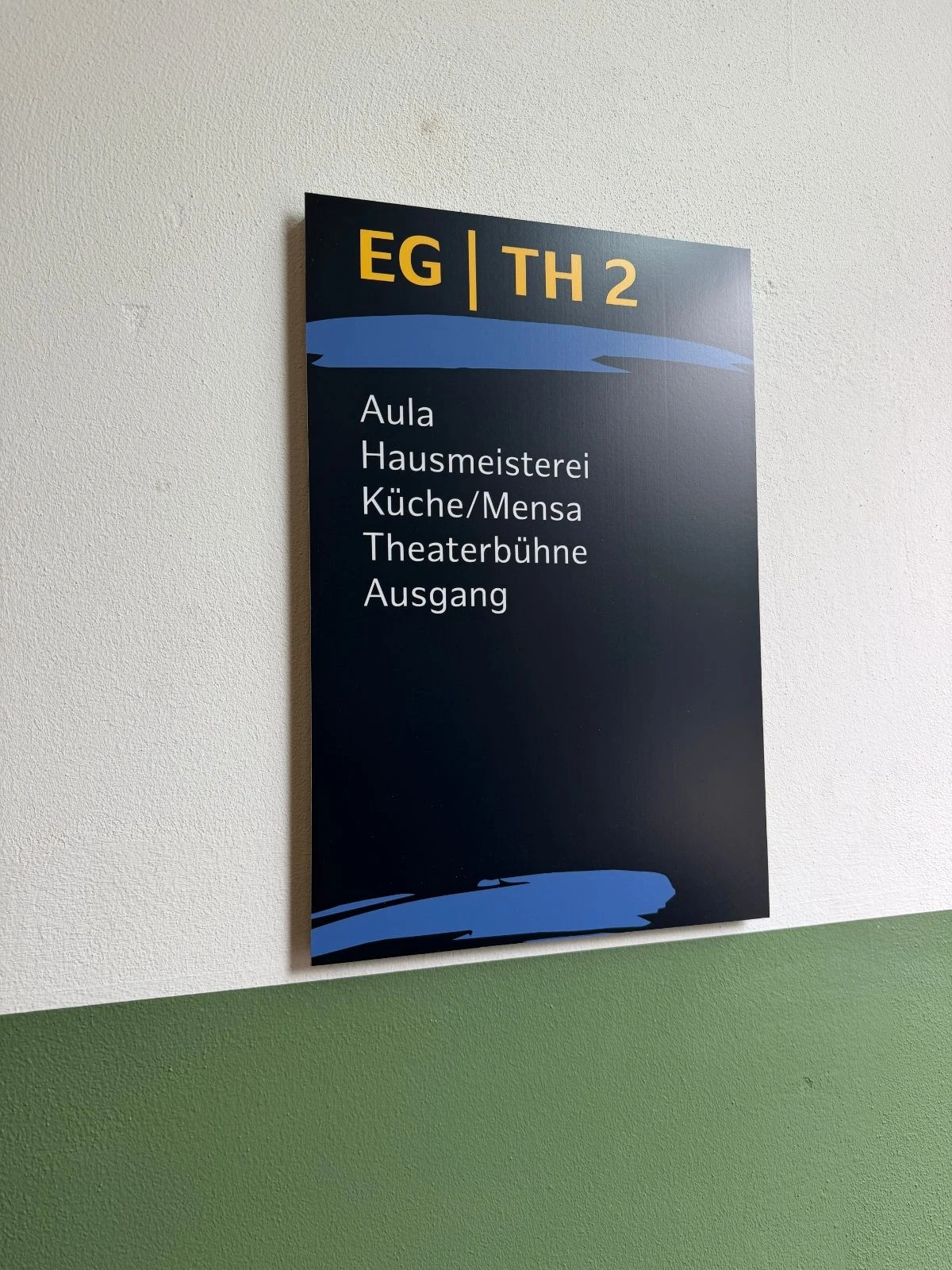 Wegweiser mit der gelben Überschrift EG | TH 2 und den Zielen Aula, Hausmeisterei, Küche/Mensa, Theaterbühne, Ausgang