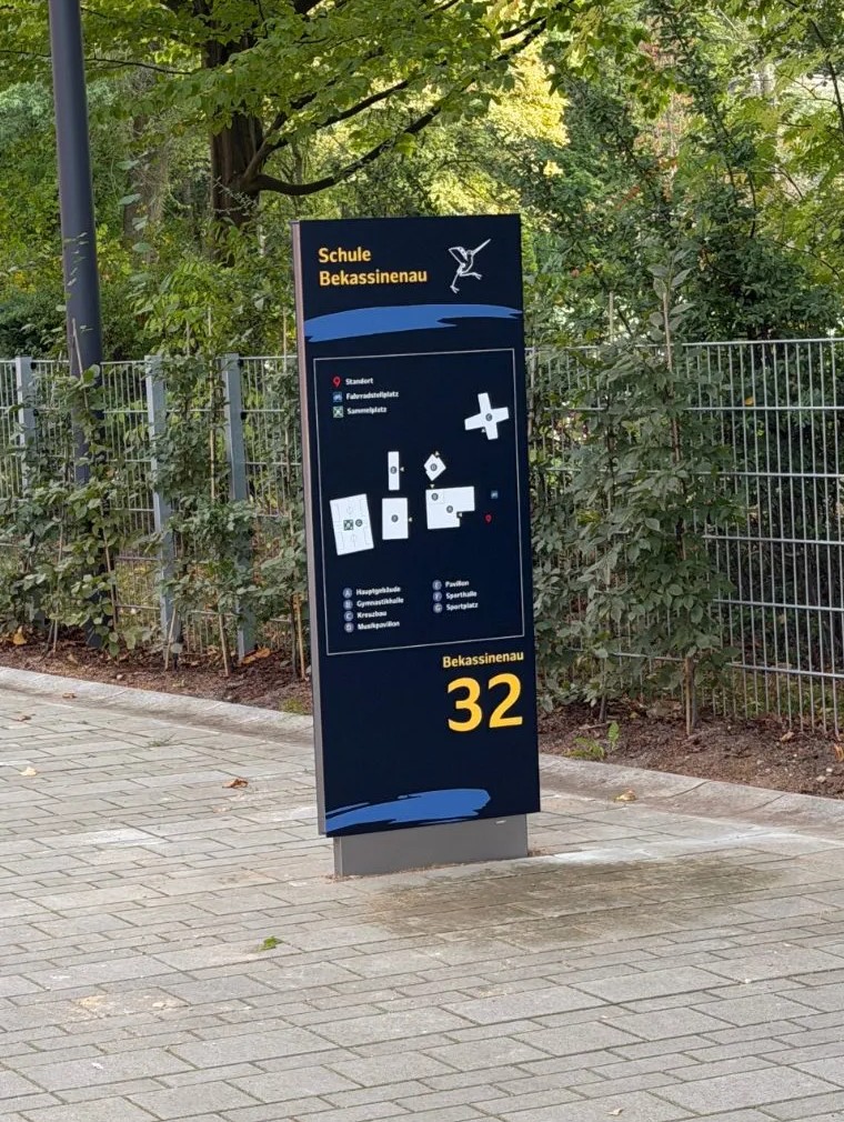 Der neue Monolith am gepflasterten Weg vor einem Zaun mit Bäumen: dunkelblaue Stele mit Schulname und Bekassine, Lageplan der Gebäude A bis G, darunter groß Bekassinenau 32 in Gelb