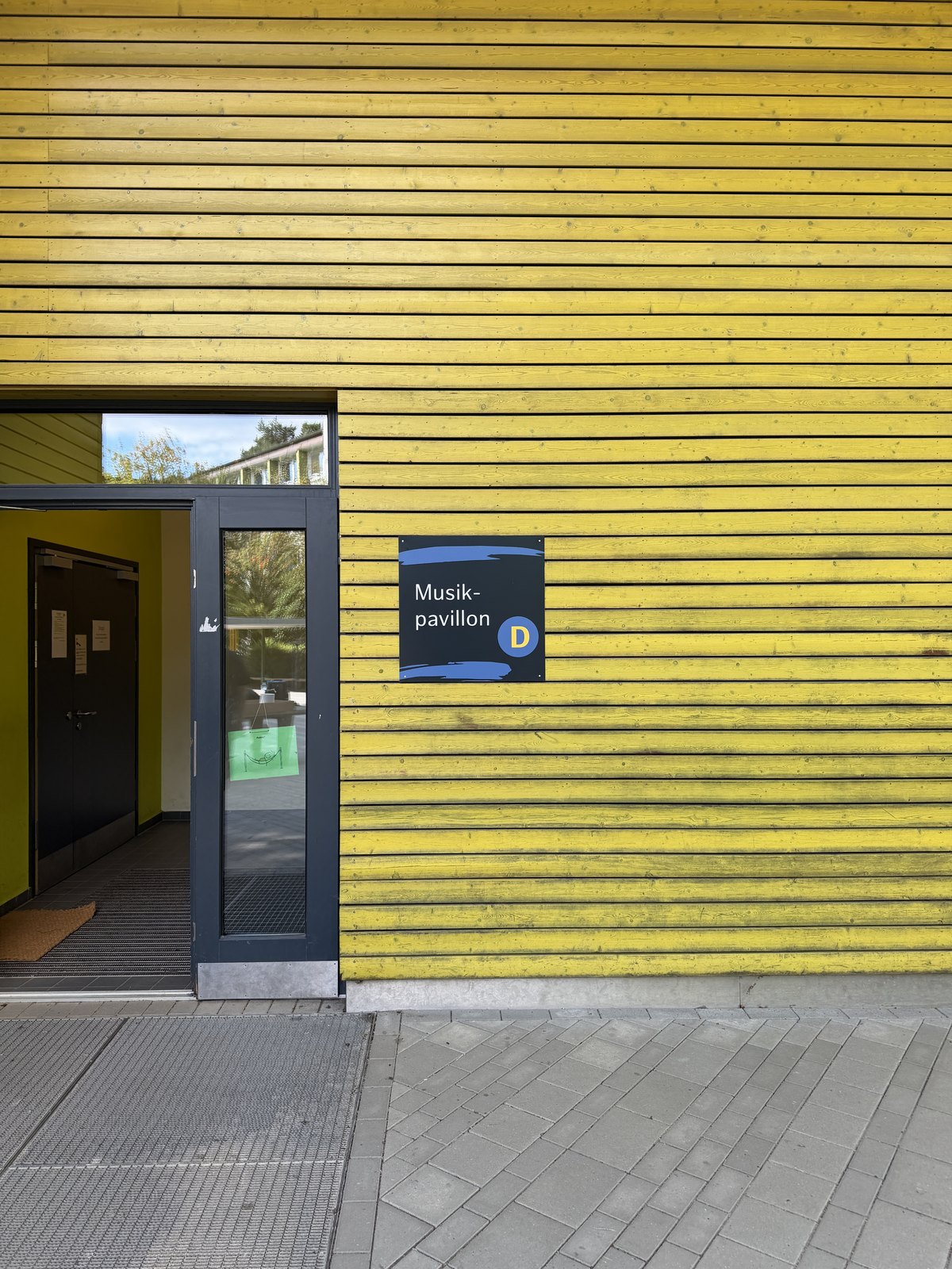 Gelbe Holzfassade mit Eingangstür, daneben ein dunkelblaues Schild mit der Aufschrift Musikpavillon und dem Buchstaben D