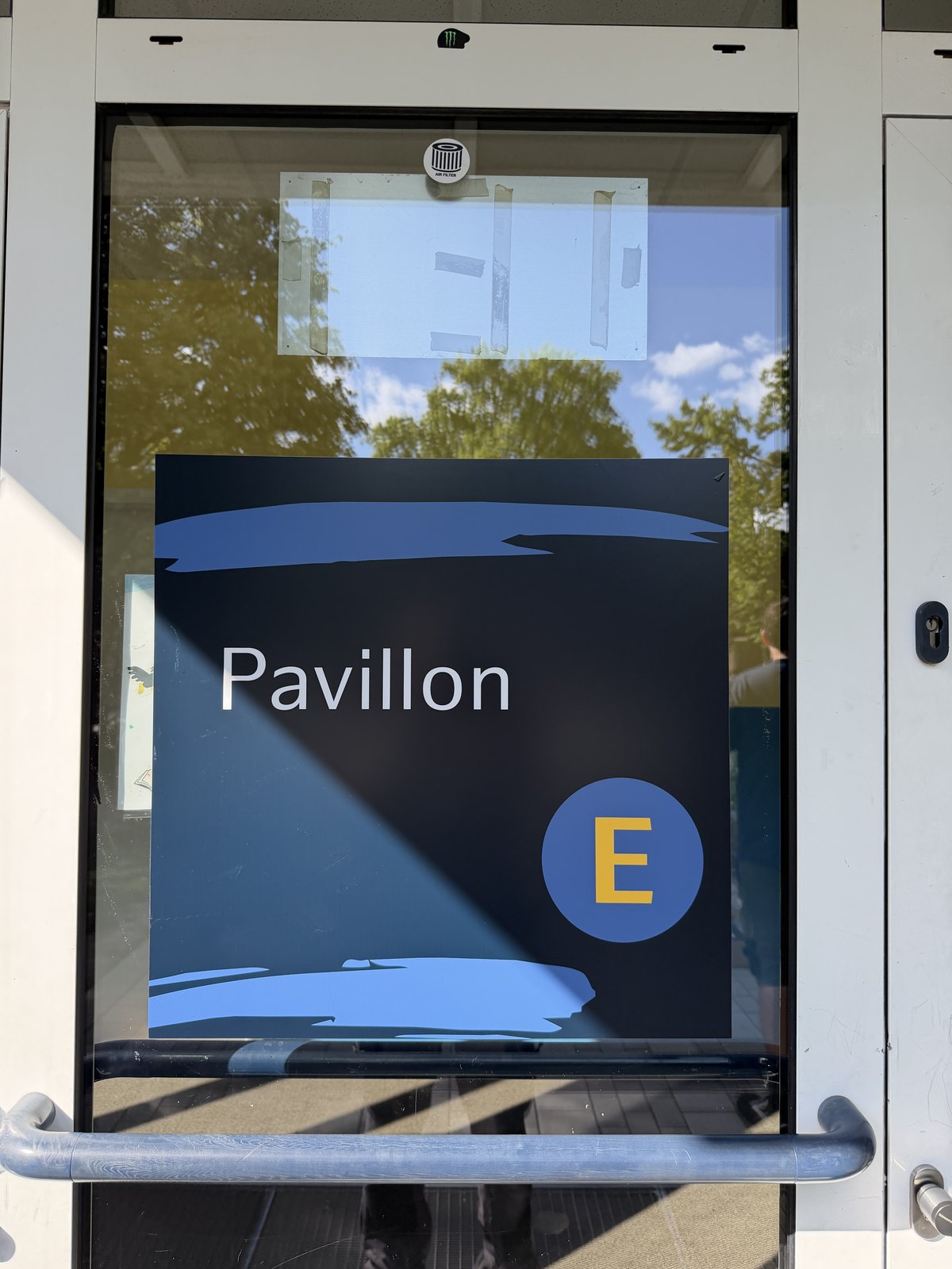 Dunkelblaues Schild als Folie auf einer Glastür mit der Aufschrift Pavillon und einem großen gelben E im blauen Kreis