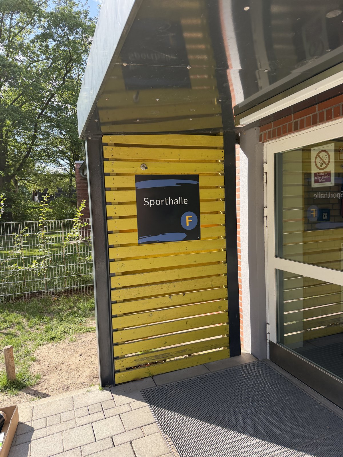 Gelbe Holzlattenfassade neben einer Tür, darauf ein dunkelblaues Schild mit der Aufschrift Sporthalle und dem Buchstaben F