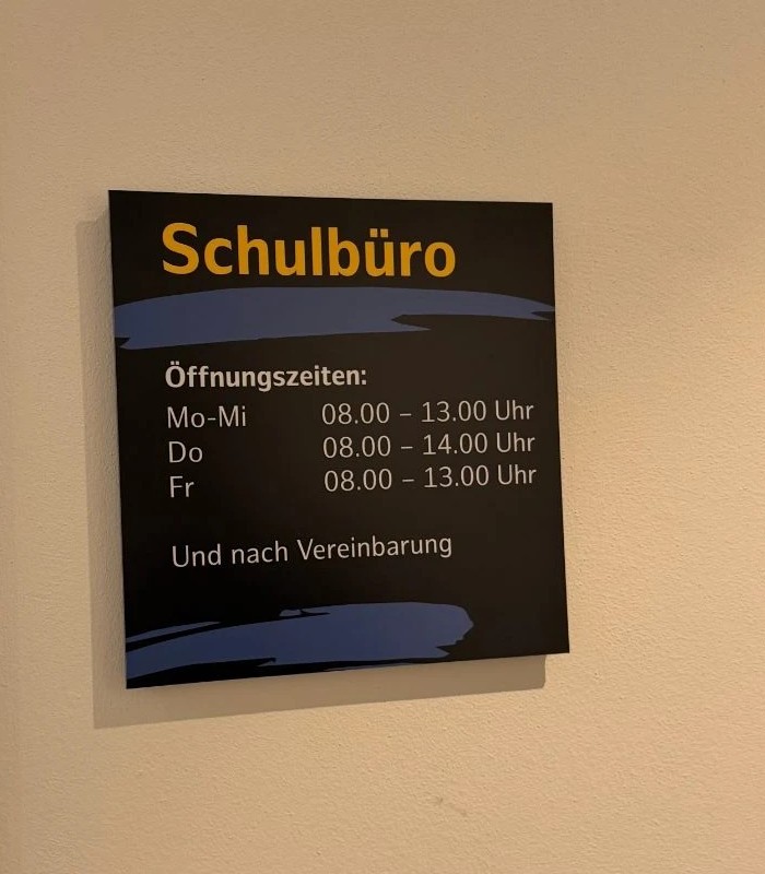 Dunkelblaues Infoschild mit der gelben Aufschrift Schulbüro und den Öffnungszeiten Mo bis Mi 8 bis 13 Uhr, Do 8 bis 14 Uhr, Fr 8 bis 13 Uhr