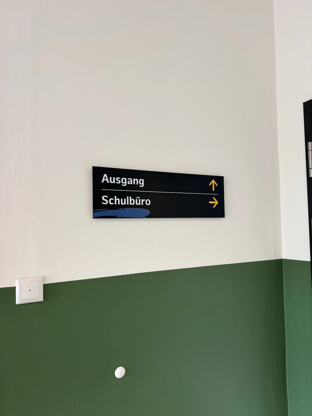 Schmaler dunkelblauer Wegweiser über einer Tür mit den Zielen Ausgang, Pfeil nach oben, und Schulbüro, Pfeil nach rechts