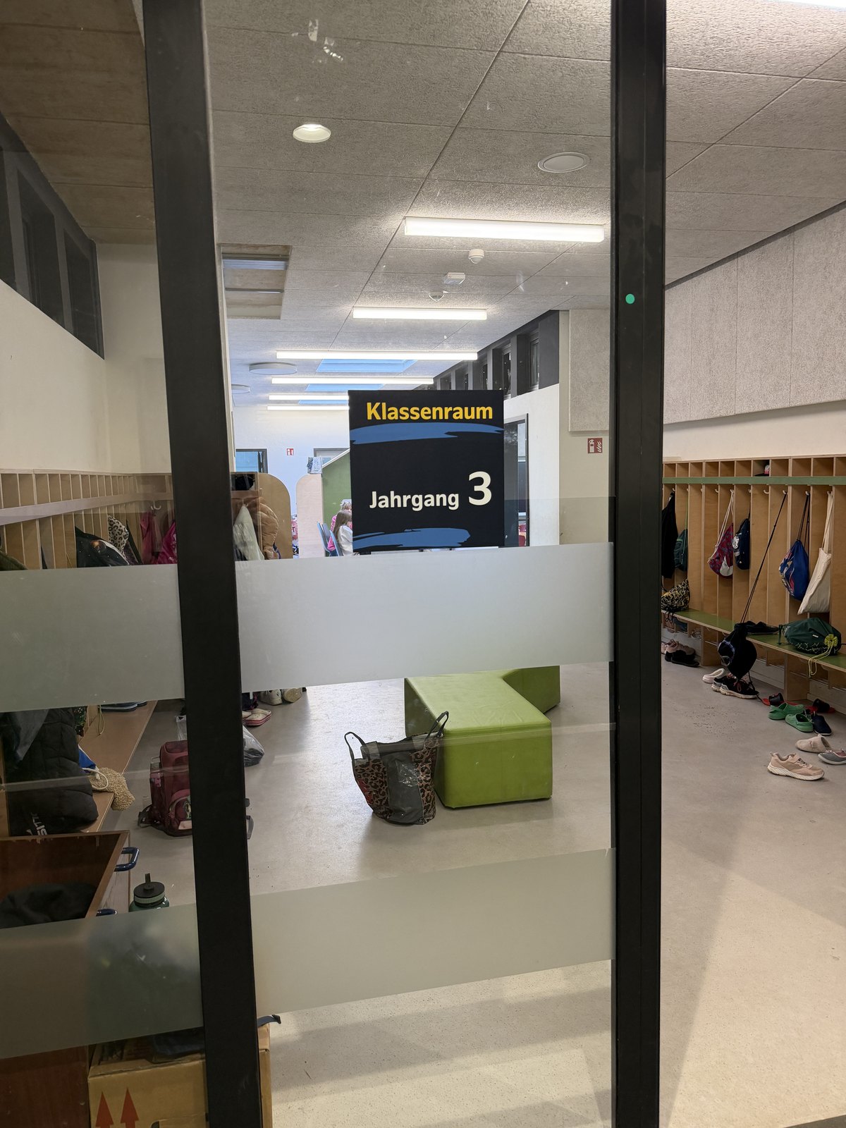Schild Klassenraum Jahrgang 3 hinter einer Glasscheibe, dahinter Garderobenhaken und Blick in den Klassenraum