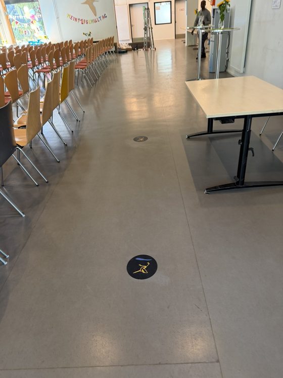 Mensa mit Stuhlreihen, auf dem hellen Boden eine runde dunkle Bodenfolie mit gelber Bekassine
