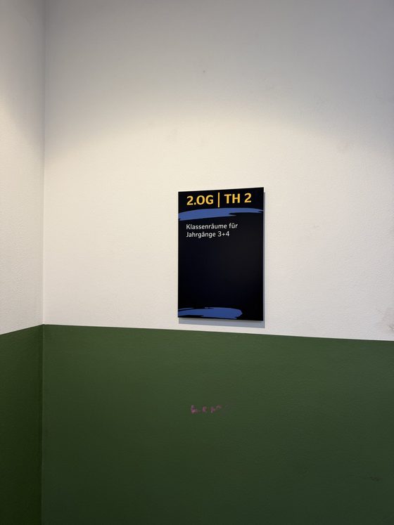 Wegweiser an weißer Wand mit der gelben Überschrift 2.OG | TH 2 und dem Ziel Klassenräume für Jahrgänge 3 und 4