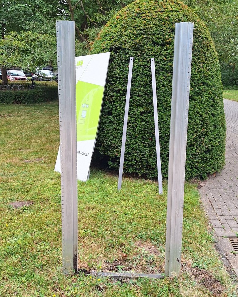 Zwei verzinkte Stahlpfosten stehen ohne Schild im Rasen, dahinter lehnt ein abgenommenes Monolith-Paneel an einer Hecke.