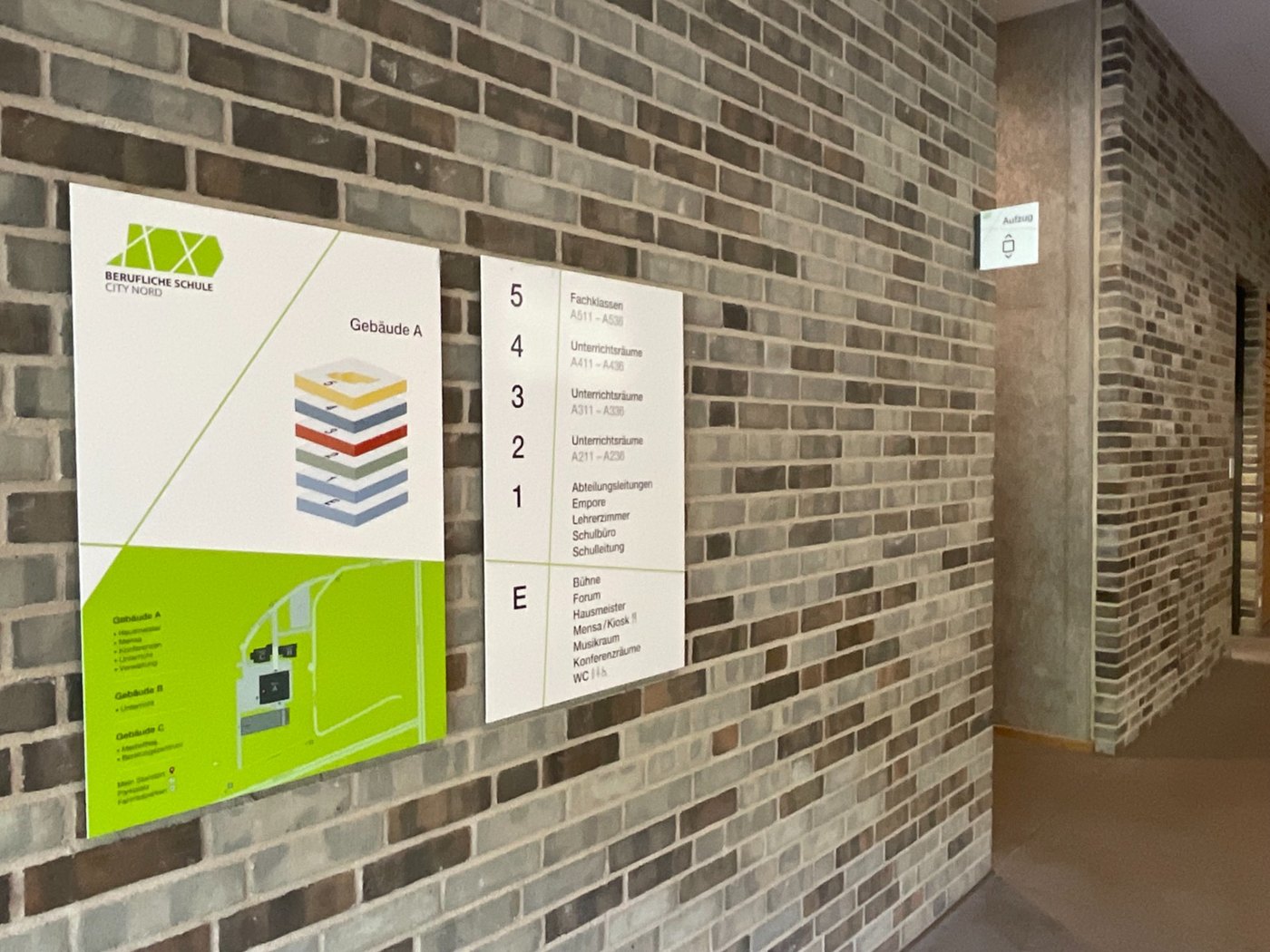 Etagenübersicht für Gebäude A an einer Klinkerwand: links ein Gebäudeschnitt mit farbig abgesetzten Ebenen über einem Campus-Lageplan, rechts die Raumgruppen je Geschoss von E bis 5.