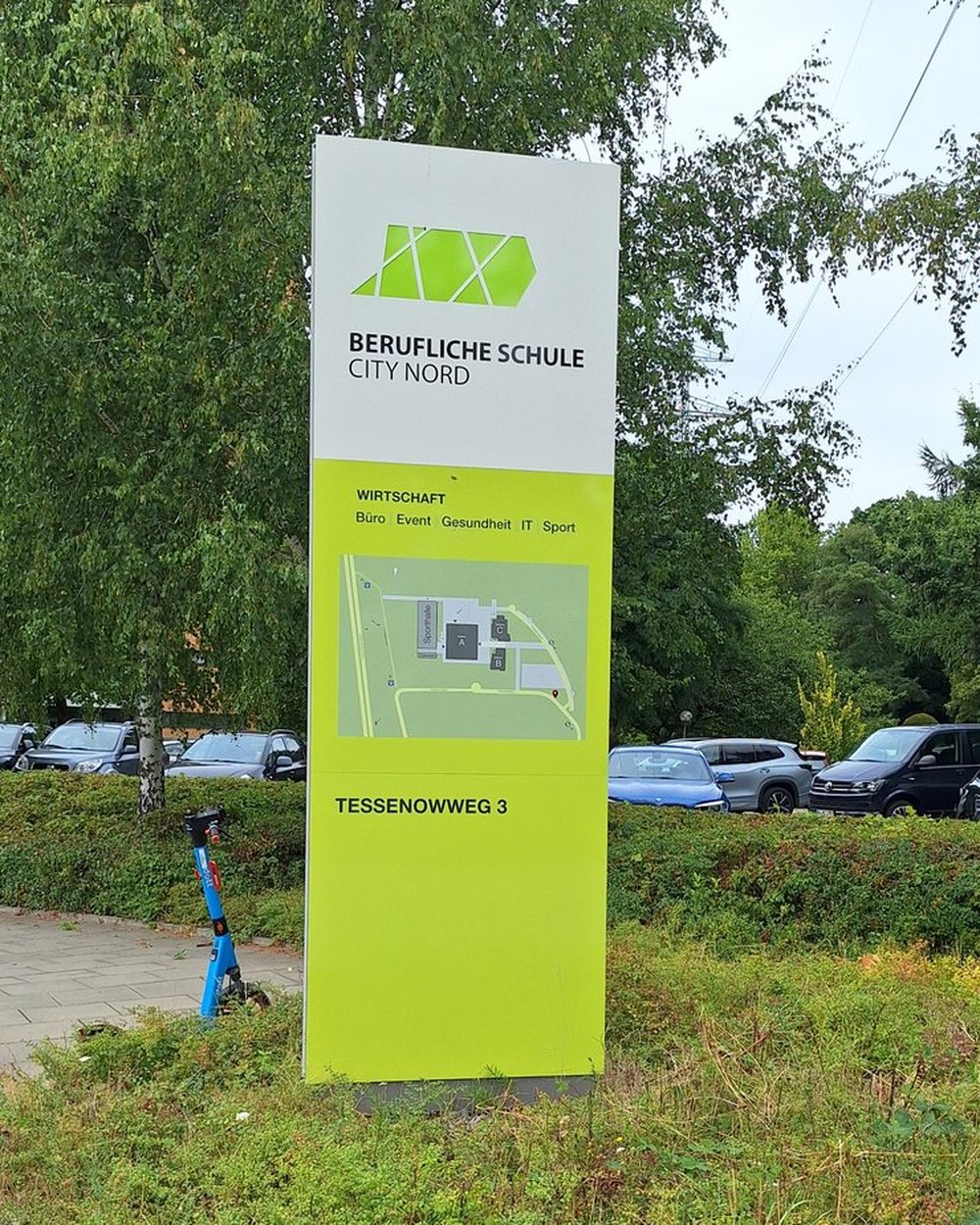 Monolith der Beruflichen Schule City Nord im bisherigen Layout am Weg, dahinter Parkplatz und Bäume.