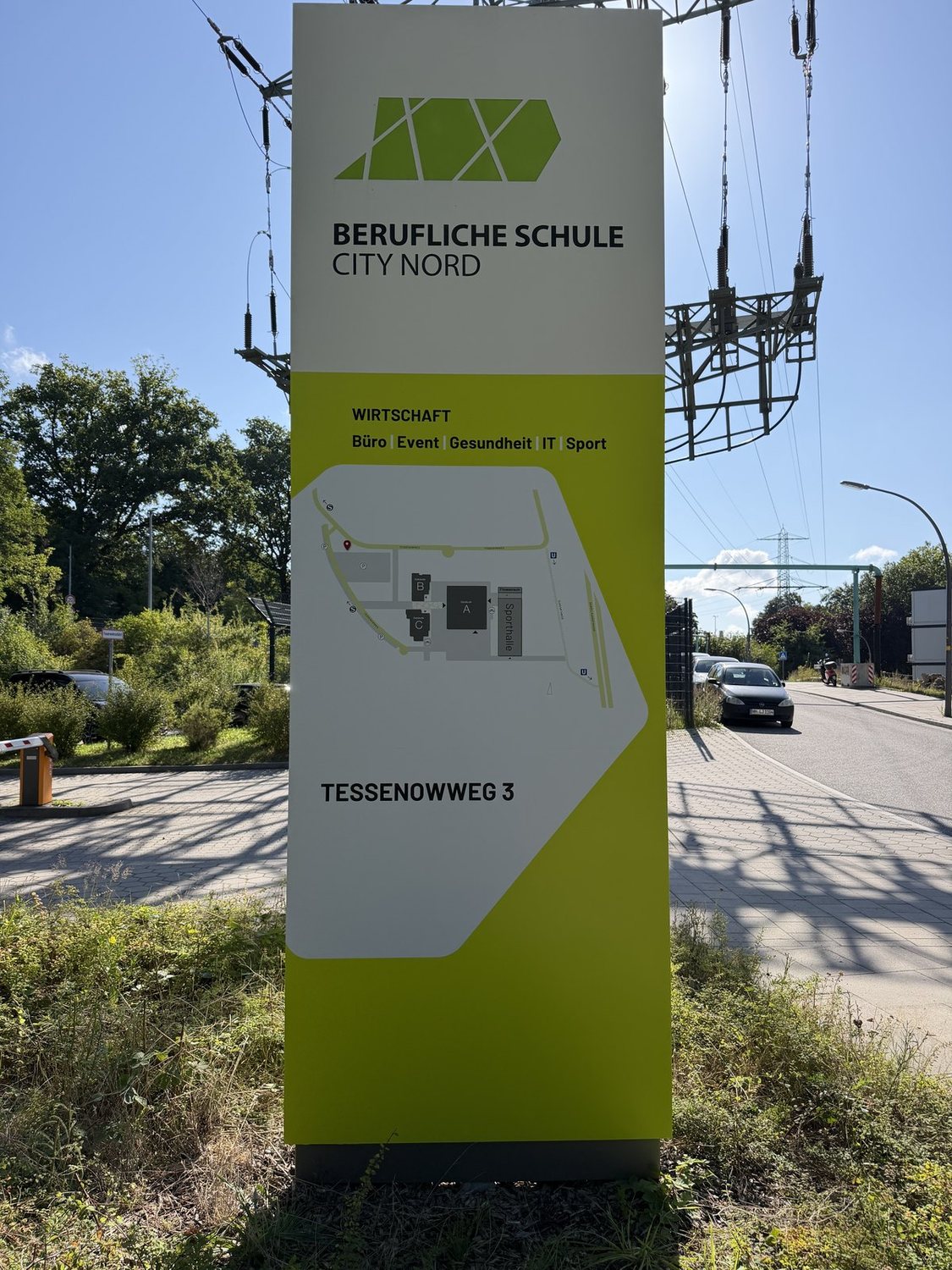 Schlanker, aluminiumgerahmter Monolith mit diagonalem Farbschnitt an der Zufahrt zur Schule, dahinter Straße und Bäume.