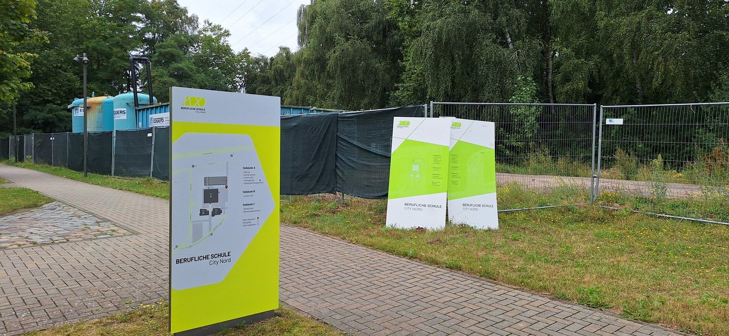 Breiter Monolith mit Campus-Lageplan und Gebäudeverzeichnis am Weg, rechts lehnen zwei abgenommene Paneele an einem Bauzaun.