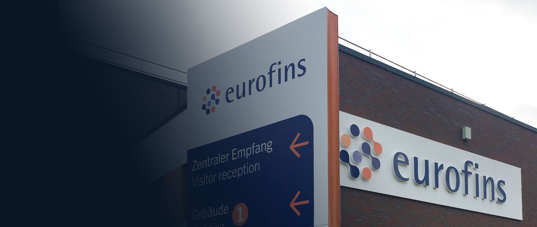 Eurofins-Gebäude mit Firmenschriftzug an der Klinkerfassade, davor ein Monolith des Außenleitsystems mit dem Ziel Zentraler Empfang