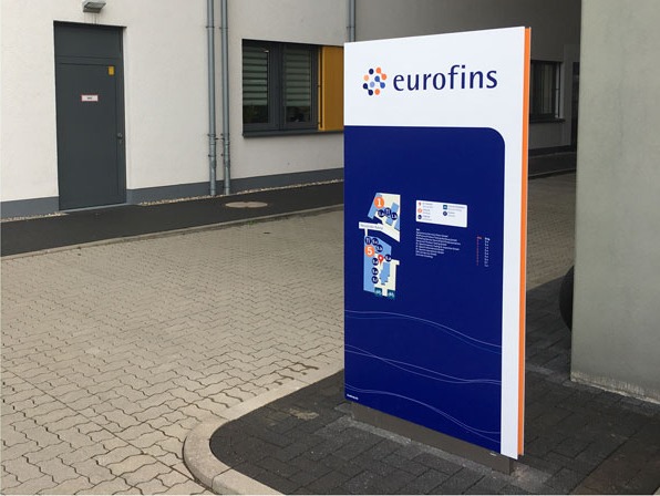 Blauer Monolith des Außenleitsystems mit Eurofins-Logo vor einem Gebäudeeingang