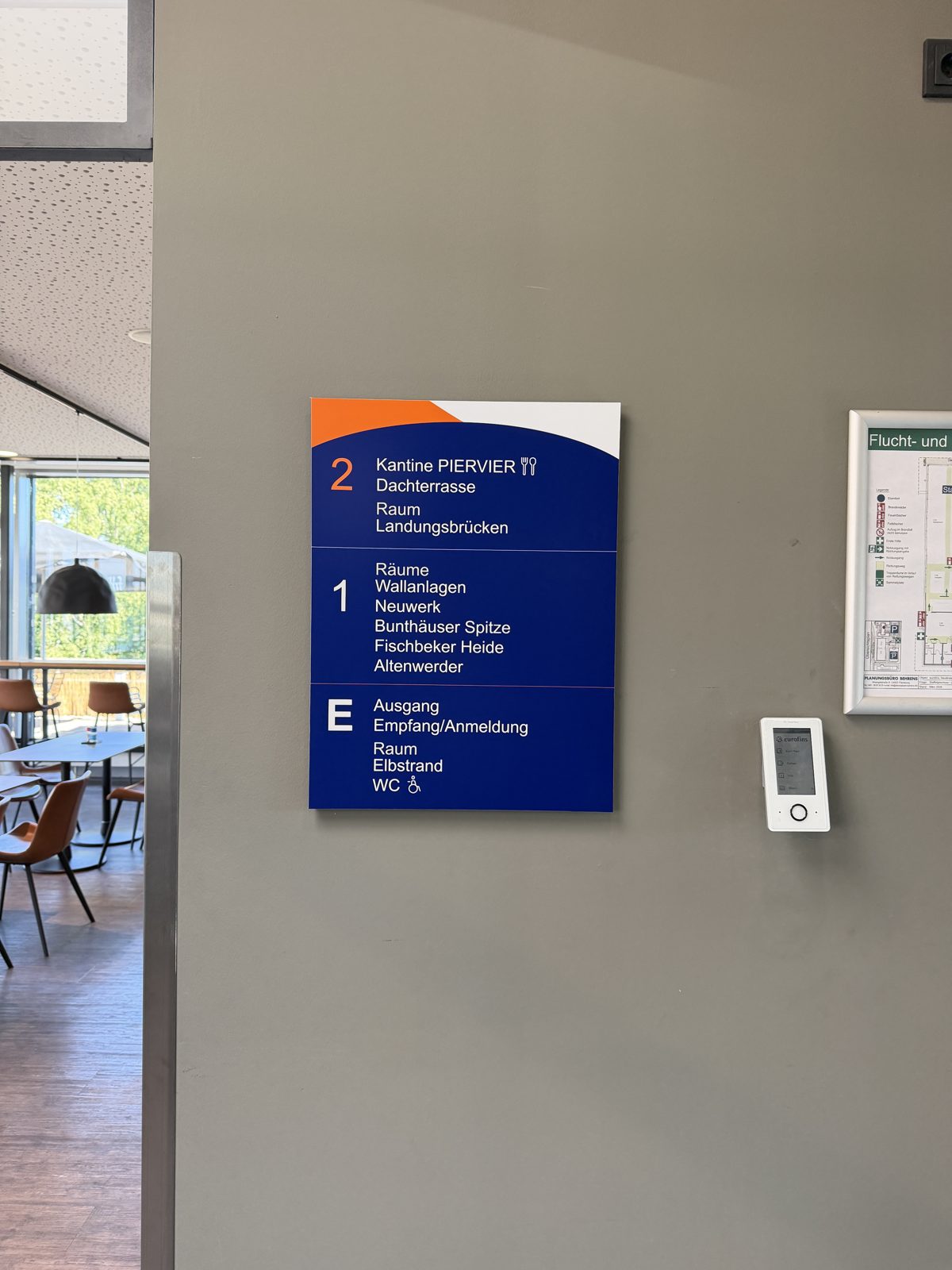 Geschosswegweiser an einer grauen Wand neben dem Kantinenbereich, die Ziffer 2 in Orange