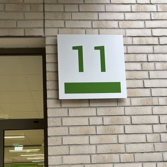 Weißes Schild mit grüner 11 und grünem Balken an einer hellen Klinkerfassade neben einer Glastür