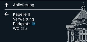 Wegweiser 6, Vorderseite: Pfeil ↑ Anlieferung; Pfeil ← Kapelle II, Verwaltung, Parkplatz, WC