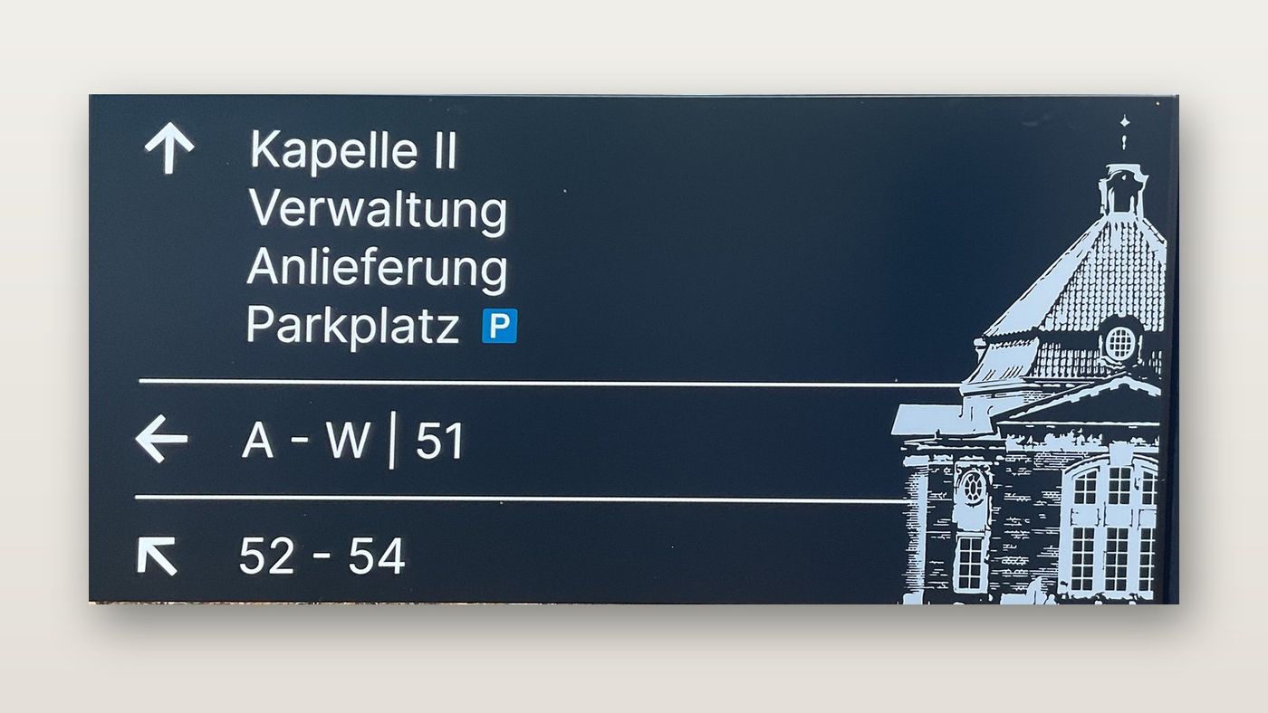 Wegweiser in Schwarzblau, freigestellt: Pfeil nach oben zu Kapelle II, Verwaltung, Anlieferung und Parkplatz, Pfeil nach links zu A bis W und 51, Schrägpfeil zu 52 bis 54, rechts die Zeichnung einer Kapelle