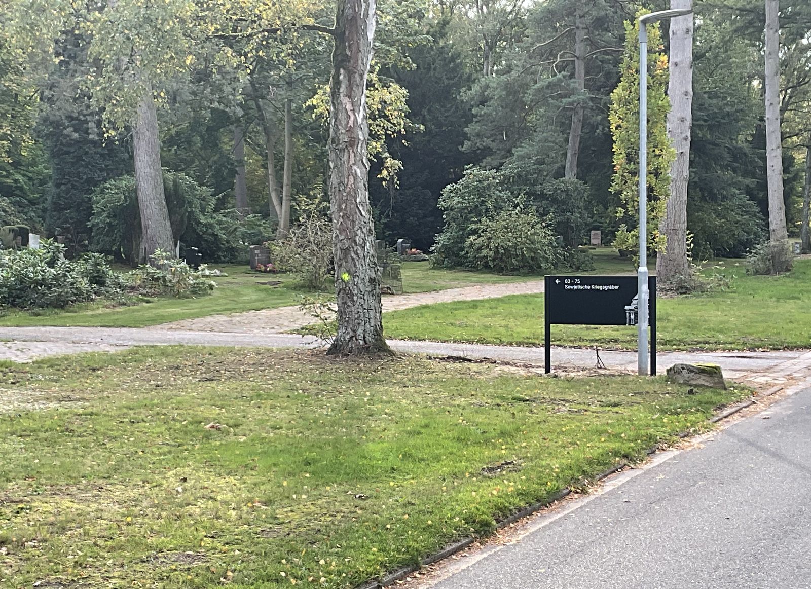 Parkähnlicher Friedhof mit hohen Bäumen und Rasen, an der Wegekreuzung ein schwarzblauer Wegweiser nach links zu den Feldern 62 bis 75 und den sowjetischen Kriegsgräbern