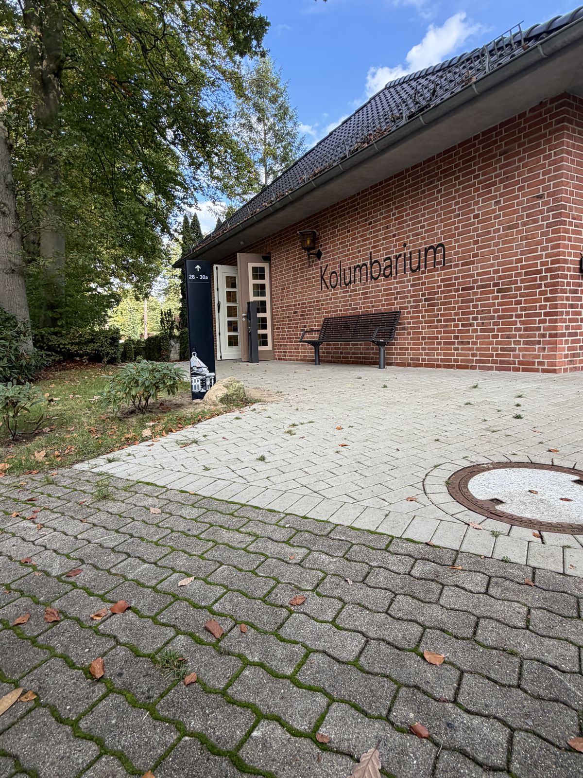 Backsteinbau mit dem Schriftzug Kolumbarium an der Fassade, davor eine gepflasterte Fläche und eine Bank