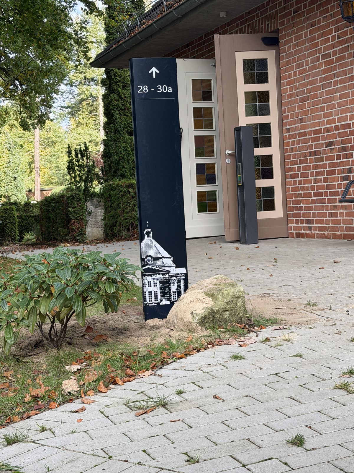 Schwarzblaue Stele vor dem Eingang eines Backsteinbaus mit Pfeil nach oben zu 28 bis 30a